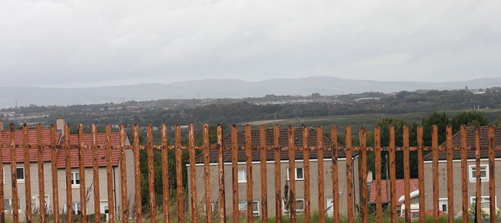 a rusty railing, beyond which is a sparawling urban skyline and a view out to hills. A cloudy sky.