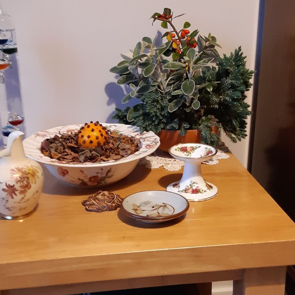 table with pot pourri, a tangerine stuck with cloves, jug, candlestick and bowl. Evergreens and berries behind.