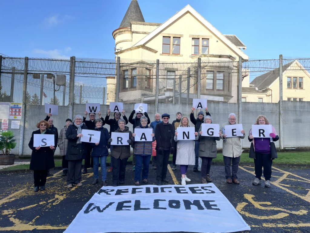 demonstration outside Dungavel detention centre, people holding up placards saying I was a Stranger, a banner on the ground saying Refugees welcome
